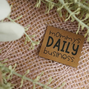 Könnte beinhalten: Ein braunes Leder-Etikett mit dem Text "mommy's DAILY business" in schwarzen Buchstaben.