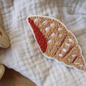 May include: A brown and white embroidered seashell patch with a red accent. The patch is made of fluffy yarn and has a white background.