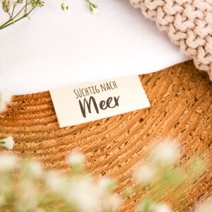 Könnte beinhalten: Nahaufnahme einer strukturierten, hellbraunen Holzoberfläche mit weißem Stoff und einem beigefarbenen Strickartikel. Ein kleines rechteckiges Etikett mit dem Text "SÜCHTIG NACH Meer" ist sichtbar. Zarte weiße Blüten sind verstreut.