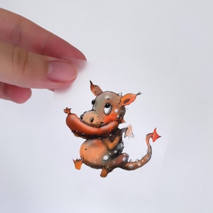 Könnte beinhalten: Eine Cartoon-Illustration eines braunen und orangefarbenen Drachen mit weißen Flecken, der eine Wurst im Maul hält. Der Drache hat einen langen Schwanz und kleine Flügel.