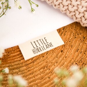 May include: A label with the text "LITTLE HURRICANE" is displayed on a textured, brown surface. A white fabric and a beige knitted item are also visible. The image is framed with delicate white flowers, creating a soft, natural aesthetic.