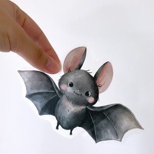 Könnte beinhalten: Eine niedliche Cartoon-Fledermaus mit schwarzem Fell, grauen Flügeln und rosa Ohren. Die Fledermaus hat große, runde Augen und eine kleine, rosa Nase.