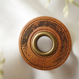 Può includere: Un bottone rotondo in legno marrone con un occhiello in ottone al centro. Il bottone ha una finitura usurata e un design a cerchi concentrici.