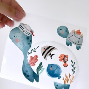Può includere: Un'illustrazione ad acquerello di una tartaruga marina blu che indossa un cappello da marinaio con un'ancora. La tartaruga è circondata da coralli, alghe e altri pesci. C'è una tartaruga marina blu più piccola sullo sfondo.