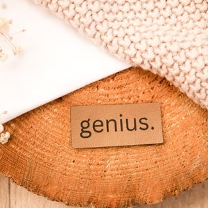 May include: A brown leather tag with the word "genius." printed in black lettering. The tag is sitting on a wooden surface with a white fabric and a knitted fabric in the background.