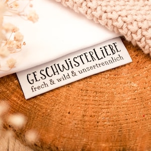 Può includere: Etichetta in tessuto bianco con testo nero che recita "Geschwisterliebe" e "frech & wild & unzertrennlich". L'etichetta è su una superficie di legno con un tessuto a maglia sullo sfondo.