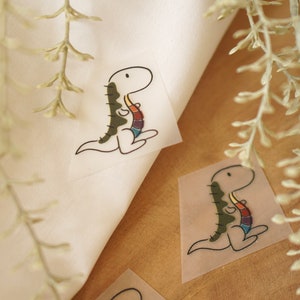 Op de afbeelding: Twee strijkbare patches met een cartoon-dinosaurus met een regenboogkleurige staart. De patches zijn op een witte stoffen achtergrond.