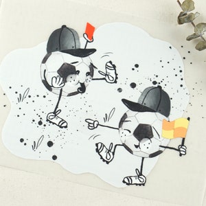 May include: Two cartoon footballs wearing baseball caps. One football is holding a red card and the other is holding a yellow and orange flag. The footballs are on a white background with black splatters.