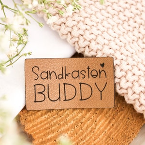 May include: A rectangular, tan leather patch with the words "Sandkasten BUDDY" in black script. A small heart is above the letter "n". The patch is on a cork surface, with a knitted blanket and white flowers in the background.