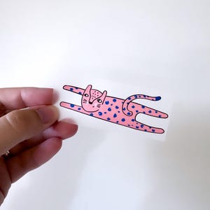 Op de afbeelding: Een roze kat met blauwe vlekken, liggend op zijn buik met zijn poten uitgestrekt. De kat heeft een eenvoudig, cartoonachtig gezicht.