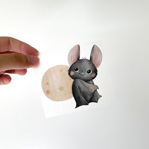 Peut inclure: Une adorable chauve-souris noire avec des oreilles roses et une lune brune derrière elle. La chauve-souris est dessinée dans un style cartoon avec de grands yeux et un sourire amical.
