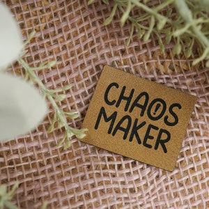 Könnte beinhalten: Ein quadratischer, hellbrauner Lederaufnäher mit den Worten "CHAOS MAKER" in schwarzen, fetten Buchstaben. Der Aufnäher befindet sich vor einem rosa gewebten Hintergrund, mit grünen und weißen dekorativen Zweigen in den Ecken.