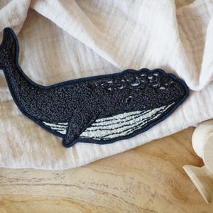 May include: A dark blue whale patch with white embroidered details and a navy blue border. The patch is lying on a cream-colored fabric, with a wooden spool in the background. The whale patch is a decorative item.