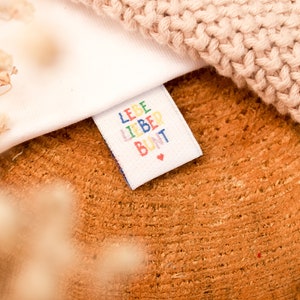 May include: A white fabric label with the text "LEBE LIEBER BUNT" in colorful letters. The label has a small red heart in the bottom right corner.