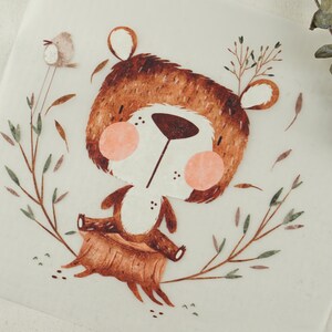 Può includere: Un orso marrone con la pancia bianca siede su un ceppo d'albero. L'orso ha il naso e le guance rosa. L'orso è circondato da foglie verdi e marroni.
