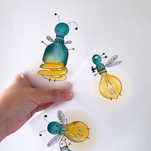 Könnte beinhalten: Drei handgemalte Glühbirnen-Illustrationen mit skurrilen Gesichtern und Flügeln. Die Glühbirnen sind in Gelb-, Grün- und Blautönen bemalt. Eine Glühbirne hält ein Glas mit Glühwürmchen.