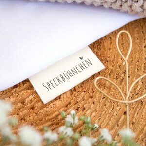Può includere: Un'etichetta in tessuto bianco con il testo "SPECKBÖHNCHEN" e un piccolo simbolo di cuore. L'etichetta è su una superficie di legno con un fiore di filo dorato.