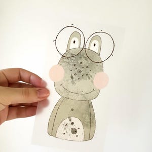 Könnte beinhalten: Eine Aquarell-Illustration eines grünen Frosches mit runden Brillen. Der Frosch hat ein breites Lächeln und rosafarbene Wangen. Die Illustration ist auf weißem Hintergrund.