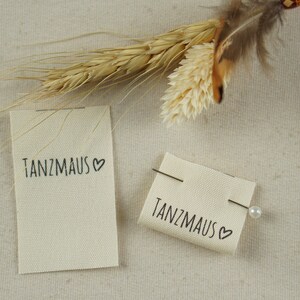 Op de afbeelding: Twee off-white stoffen labels met de tekst "Tanzmaus" en een hartsymbool in zwarte inkt. Eén label is vastgespeld met een speld. De labels liggen op een neutraal gekleurd, getextureerd oppervlak, met gedroogde tarwe en veren op de achtergrond.