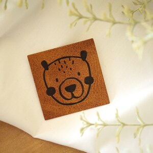 Op de afbeelding: Een bruin leren patch met een zwarte omtrek van een berenkop.