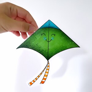 May include: A green kite sticker with a smiling face and orange and white striped tails. The kite has a blue top and is held by a hand against a white background.