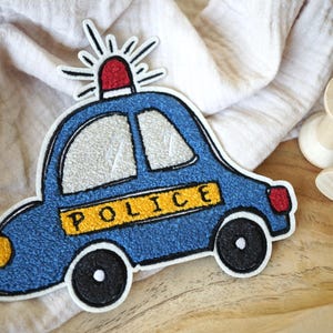 Può includere: Una toppa blu a forma di auto della polizia con la scritta "POLICE" in giallo. La toppa ha una luce rossa in cima e ruote nere. La toppa è su un tessuto di colore chiaro.
