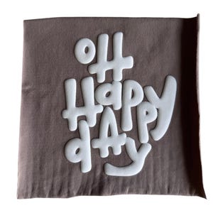 Op de afbeelding: Een bruin stoffen kussen met de woorden "Oh Happy Day" in witte, opgevulde letters.