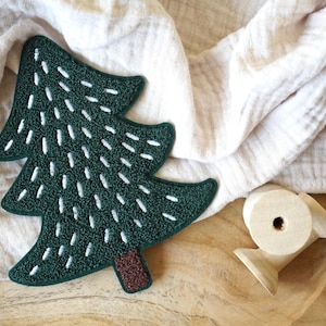 Könnte beinhalten: Ein grüner Filz-Weihnachtsbaum-Anhänger mit weißen Nähten. Der Baum liegt auf einer Holzoberfläche mit einem weißen Tuch im Hintergrund.