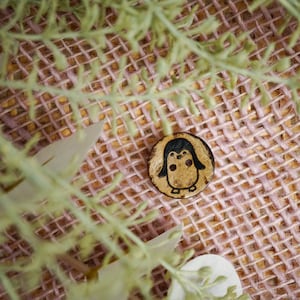 Peut inclure: Un bouton en bois avec un motif de pingouin. Le pingouin est brun avec des yeux noirs et un bec noir. Le bouton est sur un fond de toile de jute rose.