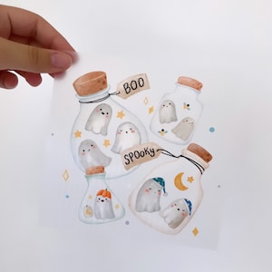 Può includere: Cinque barattoli di vetro con tappi di sughero contengono simpatici fantasmi dei cartoni animati. I barattoli hanno etichette scritte a mano che dicono "Boo" e "Spooky".