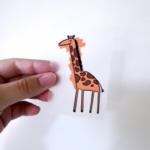 Op de afbeelding: Een grillige sticker met een girafillustratie. De giraf heeft een lange nek, bruine vlekken en een zwarte manen. De sticker wordt tegen een witte achtergrond gehouden.