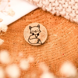 Peut inclure: Un bouton en bois avec un motif de renard. Le bouton est brun et a un contour noir. Le renard est tourné vers la droite et a un ventre blanc.
