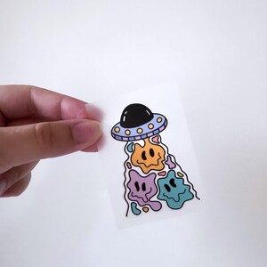 Könnte beinhalten: Ein Aufkleber mit einem Cartoon-UFO mit drei tropfenden Smiley-Gesichtern in Orange, Lila und Blau.