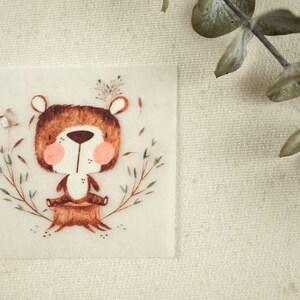 Könnte beinhalten: Ein brauner Bär mit rosa Wangen sitzt auf einem Baumstumpf, umgeben von grünen Blättern. Der Bär hat einen weißen Bauch und trägt eine braune Weste. Der Hintergrund ist weiß.