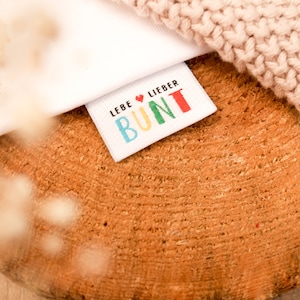 Peut inclure: Une étiquette en tissu blanc avec le texte "LEBE ♥ LIEBER BUNT" en lettres colorées. L'étiquette est sur une surface en bois avec un tissu tricoté en arrière-plan.