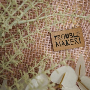 May include: A brown leather patch with the text "TROUBLE MAKER!" in black lettering. The patch is on a pink and brown hessian background.