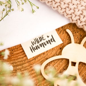 Può includere: Un'etichetta in tessuto bianco con il testo "WILDE ♥ HUMMEL" in corsivo nero. L'etichetta è su una superficie di legno con un ritaglio di legno a forma di ape.