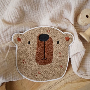 Può includere: Un toppa ricamata a forma di testa di orso marrone e beige con naso marrone e occhi neri. La toppa è su uno sfondo di tessuto beige.