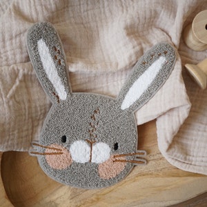 May include: A grey felt bunny head with white ears and pink cheeks. The bunny is on a wooden surface with a beige fabric in the background.
