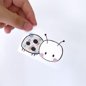 Könnte beinhalten: Ein weißer Aufkleber mit einem Cartoon-Marienkäfer-Design. Der Marienkäfer hat einen weißen Kopf mit rosa Wangen und einem schwarzen Umriss. Der Körper ist eine Mischung aus Farben und Mustern. Der Aufkleber wird von einer Hand gehalten.