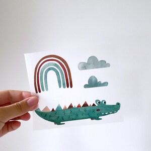May include: A green alligator with brown teeth and red eyes, a rainbow with red, orange, and blue stripes, and two grey clouds on a white background.