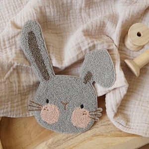 May include: A grey felt bunny rabbit with brown ears and pink cheeks. The bunny is on a wooden surface with a beige fabric background.