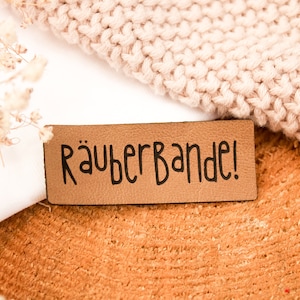 Op de afbeelding: Een bruin leren label met de tekst "Räuberbande!" in zwarte letters.
