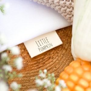 Könnte beinhalten: Ein weißes Stofflabel mit dem Text "LITTLE PUMPKIN" in schwarzer Schrift. Das Label befindet sich auf einer Holzoberfläche mit einer weißen Blume.