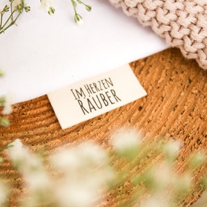 Op de afbeelding: Een wit stoffen label met zwarte tekst die "Im Herzen Räuber" luidt, op een houten oppervlak met witte bloemen.