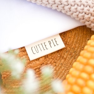 Può includere: Un'etichetta in tessuto bianco con il testo "CUTTIE PIE" stampato in nero su una superficie in legno con un tessuto a maglia e una pannocchia di mais sullo sfondo.