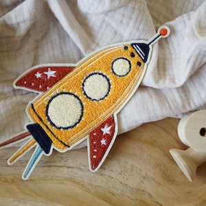 Peut inclure: Un patch brodé en forme de fusée jaune et rouge avec des étoiles blanches et trois cercles blancs. La fusée a une queue bleue et noire avec trois flammes.