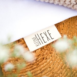 Könnte beinhalten: Nahaufnahme eines weißen Stofflabels mit schwarzem Text "kleine HEXE" auf einer Holzoberfläche.