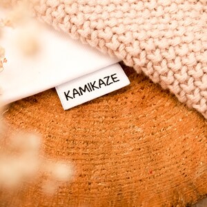 May include: A white fabric label with the word "KAMIKAZE" printed in black lettering. The label is lying on a wooden surface with a knitted fabric on top of it.