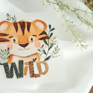 Könnte beinhalten: Eine Aquarell-Illustration eines niedlichen Tigers mit braunen Streifen und einer rosa Nase. Der Tiger ist von grünen Blättern umgeben und das Wort "WILD" ist darunter in Braun, Schwarz und Weiß geschrieben.